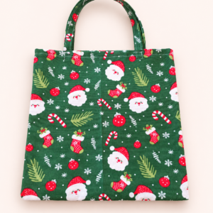 Grüne Stofftasche mit Weihnachtsmannköpfen, roten Weihnachtsstrümpfen, Zuckerstangen, Kugeln und weißen Schneeflocken vor hellem Hintergrund.