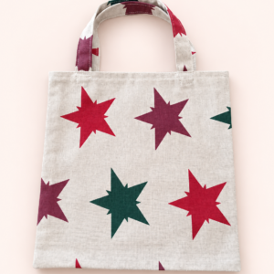 Helle Stofftasche mit zwei Henkeln und großen roten, bordeauxfarbenen und grünen Sternen vor hellem Hintergrund.